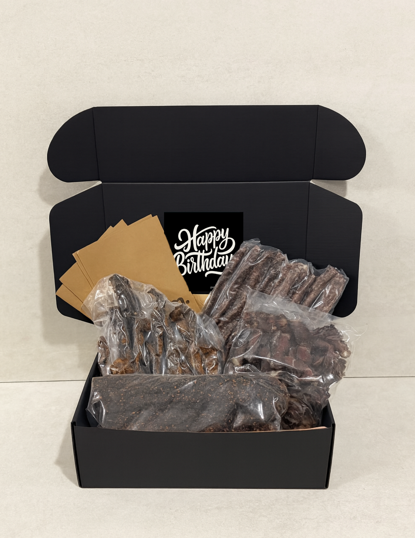 Black gift box with 'Happy Birthday' message containing biltong and droewors on a light gray background