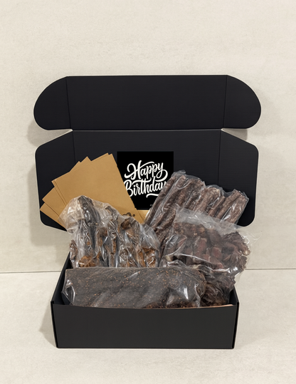 Black gift box with 'Happy Birthday' message containing biltong and droewors on a light gray background