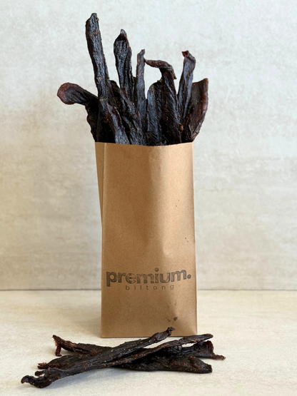 Dried meat sticks stokkies jerky in a brown paper bag labeled 'premium biltong' on a light background
