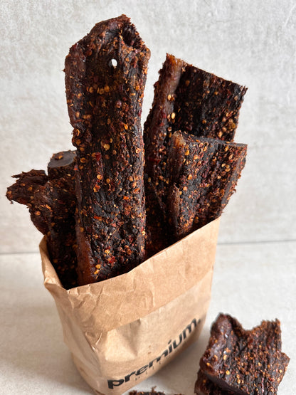 A paper bag filled with whole pieces of chilli biltong, a traditional South African dried meat snack, seasoned with salt, pepper, chilli and coriander.