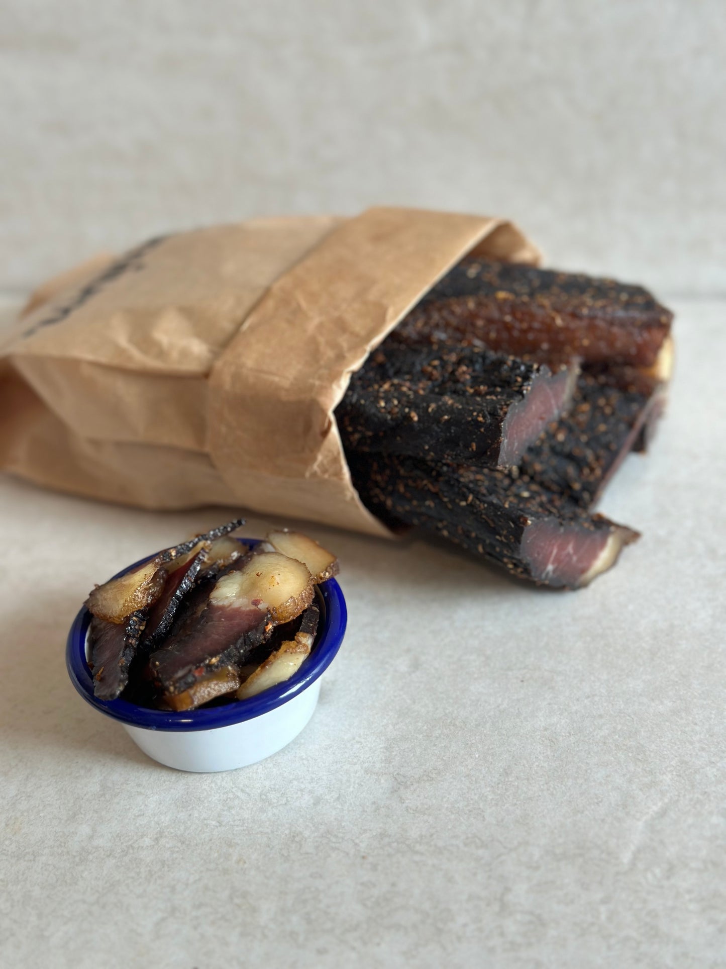 Sliced Fatty Biltong in a blue container with a brown paper bag full of whole biltong slabs on a light gray background
